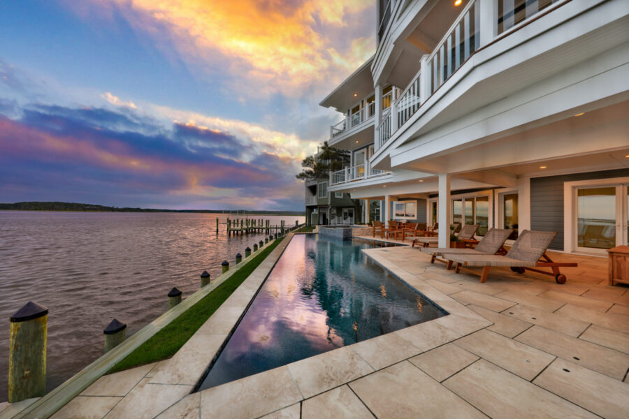 Modern waterfront house with multiple balconies, an infinity pool, and lounge chairs overlooking a calm body of water at sunset—perfect for outdoor living and relaxing in style.