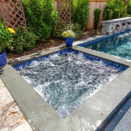 A backyard hot tub with bubbling water is adjacent to a luxury pool, surrounded by green plants and potted flowers.