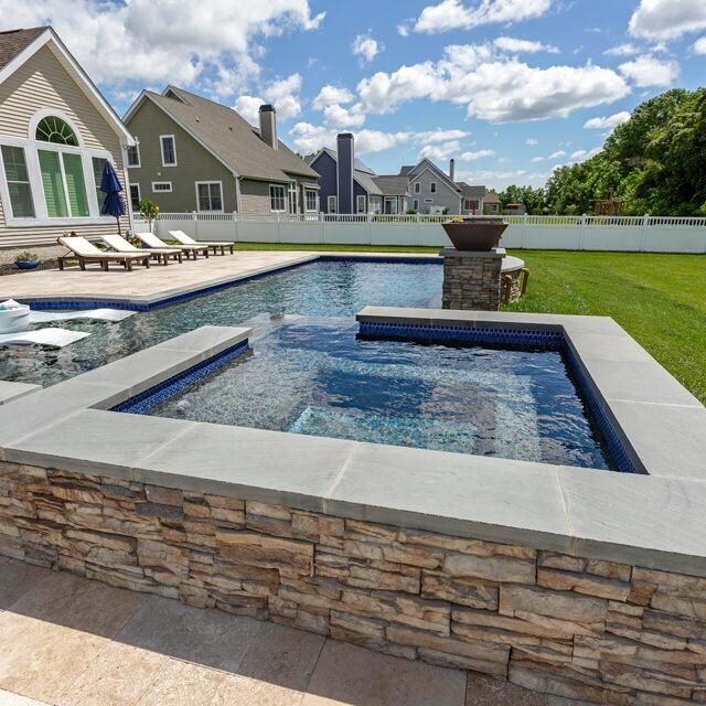 Modern backyard featuring a rectangular fiberglass pool with an attached custom spa, stone edging, lounge chairs, and well-kept lawn—perfect for outdoor living, bordered by houses and a white fence.