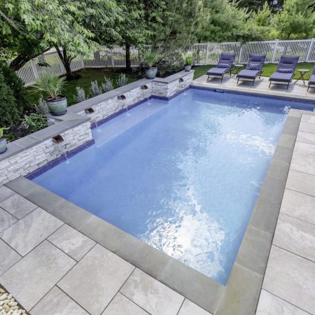 Rectangular backyard inground pool with light stone tile deck, four water features, potted plants, and six lounge chairs along one side, surrounded by trees and a white fence. Perfect for relaxing or entertaining near future outdoor kitchens.
