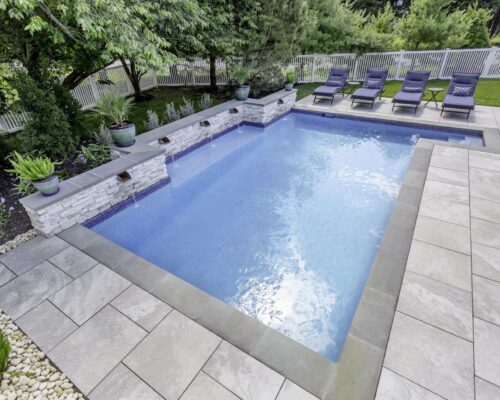Rectangular backyard inground pool with light stone tile deck, four water features, potted plants, and six lounge chairs along one side, surrounded by trees and a white fence. Perfect for relaxing or entertaining near future outdoor kitchens.