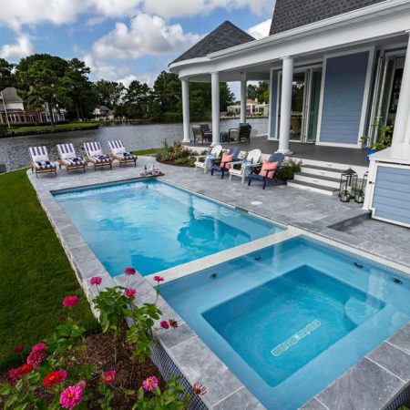 A backyard featuring a luxury inground pool and hot tub, lounge chairs, a patio seating area, and a house overlooking a lake with trees and other houses in the background.