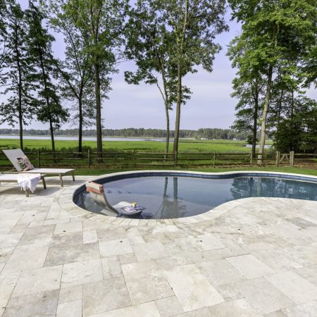 A kidney-shaped pool with two lounge chairs, a towel, and a potted plant on a stone patio creates the perfect outdoor living space, overlooking a grassy field and trees.