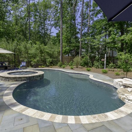 A backyard oasis featuring a fiberglass pool with built-in hot tub, stone patio, rock feature, seating area with umbrella—perfect for outdoor living and surrounded by lush trees and greenery.