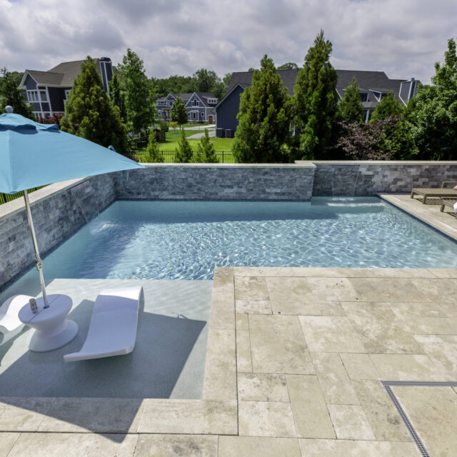 Rectangular fiberglass pool with stone tile deck, lounge chairs, and a blue umbrella—perfect for outdoor living—surrounded by trees and neighboring houses in the background.