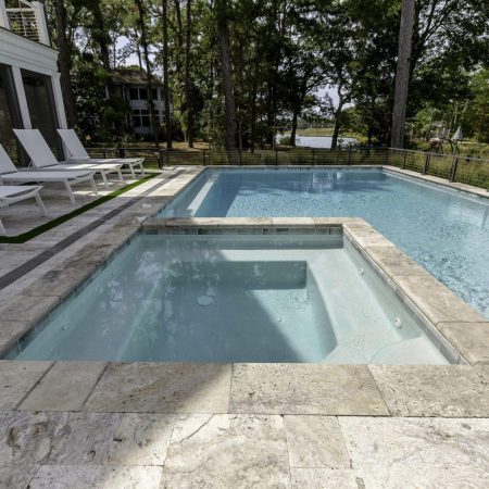 A rectangular custom spa connects to a larger inground pool on a stone patio, with several white lounge chairs lined up nearby and trees visible in the background.