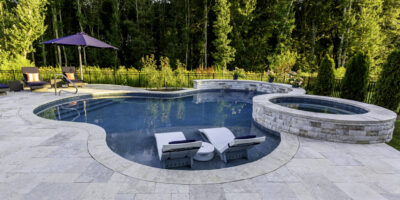 A backyard inground pool with two lounge chairs in shallow water, a circular hot tub attached, and patio seating with an umbrella, surrounded by trees and greenery, creates the perfect setting for luxury pools.