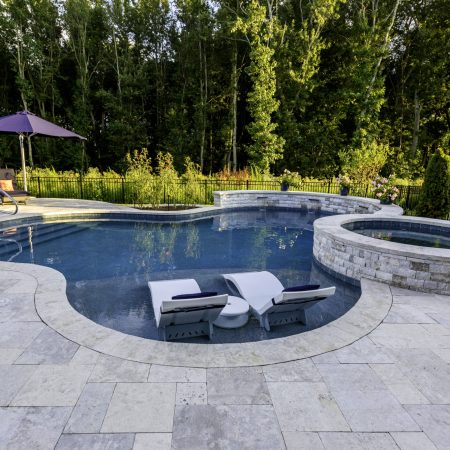 A backyard inground pool with two lounge chairs in shallow water, a circular hot tub attached, and patio seating with an umbrella, surrounded by trees and greenery, creates the perfect setting for luxury pools.