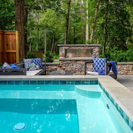 A backyard inground pool next to a stone patio with outdoor seating, a fireplace, and surrounded by trees and a wooden privacy fence—perfect for relaxing or entertaining.
