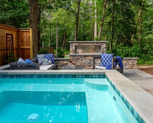 A backyard inground pool next to a stone patio with outdoor seating, a fireplace, and surrounded by trees and a wooden privacy fence—perfect for relaxing or entertaining.