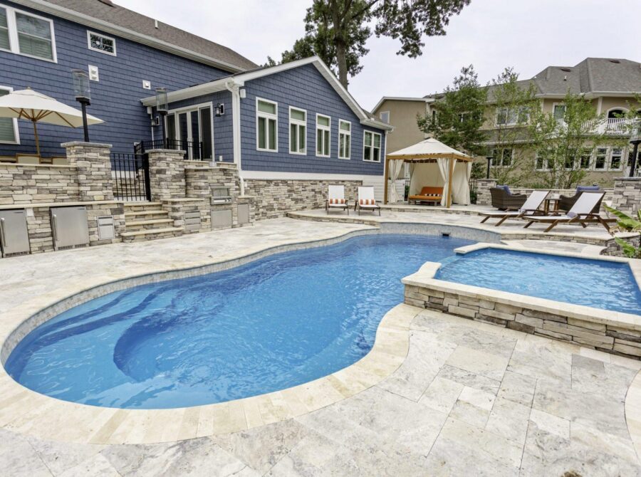 Backyard with stone patio, luxury pool, custom spa hot tub, lounge chairs, sun umbrella, and a cabana next to a blue-sided house—perfect for relaxing by the best inground pools.
