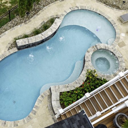 Aerial view of a kidney-shaped inground pool with an attached circular hot tub, surrounded by stone patio, lounge chairs, outdoor living spaces, and lush landscaping.