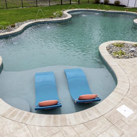 Curved inground pool with two blue loungers in shallow water, surrounded by a stone deck, small fountains, landscaping, and perfect for outdoor living.