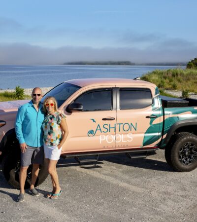 A man and woman stand in front of a pickup truck with Ashton Pools branding, parked by a beach with water and trees—perfect for discussing outdoor kitchens or upgraded outdoor living spaces.