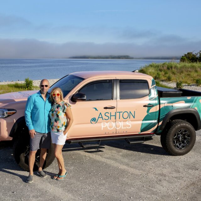 A man and woman stand in front of a pickup truck with Ashton Pools branding, parked by a beach with water and trees—perfect for discussing outdoor kitchens or upgraded outdoor living spaces.