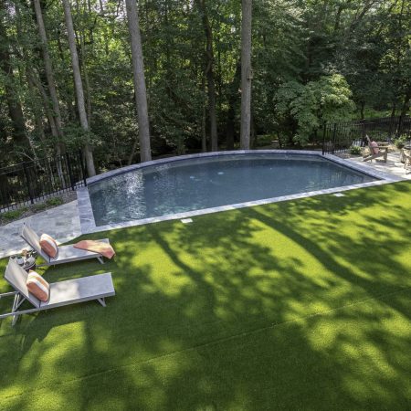 A backyard with an in-ground luxury pool, custom spa, lounge chairs on artificial grass, and a fire pit area with seating, surrounded by trees and a black metal fence—perfect for outdoor living.