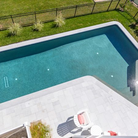 Aerial view of a modern, angular fiberglass pool with clear water, surrounded by a stone deck, grass, and black metal fence. Two white lounge chairs with red cushions are visible. Perfect for homes with custom spas or outdoor kitchens.