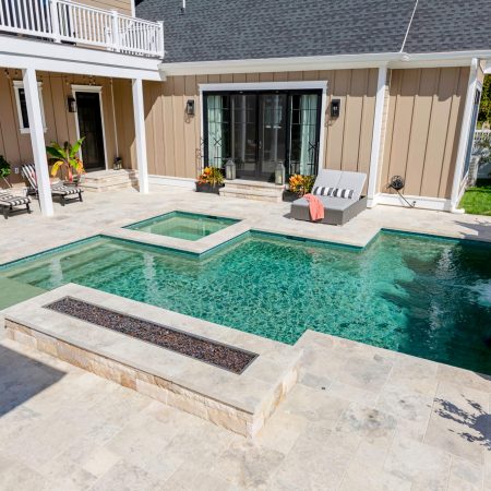 Rectangular fiberglass pool with a built-in hot tub, surrounded by stone tile, patio furniture, and potted plants—perfect for outdoor living beside a beige house with glass doors.