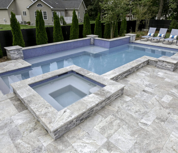 A modern backyard featuring an inground pool with a custom spa, surrounded by stone tiles, lounge chairs, and tall shrubs for privacy—perfect for outdoor living.
