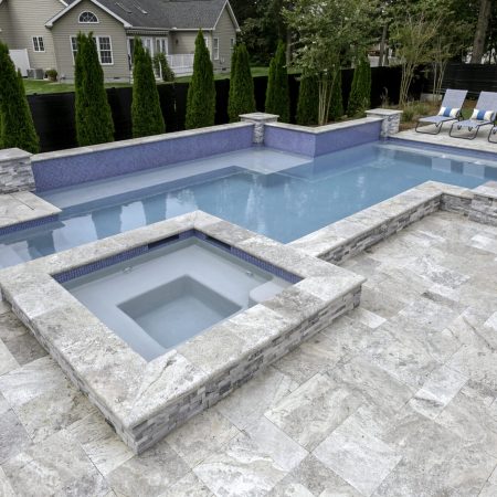A backyard with a rectangular inground swimming pool and attached hot tub, surrounded by stone tile flooring, lounge chairs, privacy trees, and designed for the ultimate outdoor living experience.