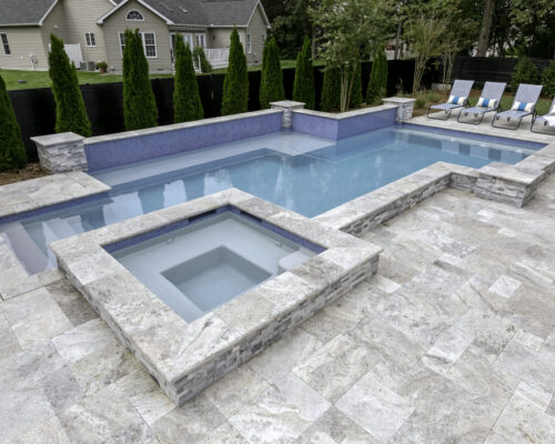 A backyard with a rectangular inground swimming pool and attached hot tub, surrounded by stone tile flooring, lounge chairs, privacy trees, and designed for the ultimate outdoor living experience.