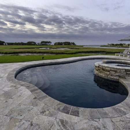 A swimming pool with an attached circular hot tub, surrounded by stone tiles, overlooks a grassy area and waterway under a cloudy sky—perfect for outdoor living. Lounge chairs and a striped umbrella are nearby for relaxing by the inground pool.