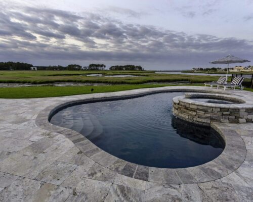 A swimming pool with an attached circular hot tub, surrounded by stone tiles, overlooks a grassy area and waterway under a cloudy sky—perfect for outdoor living. Lounge chairs and a striped umbrella are nearby for relaxing by the inground pool.