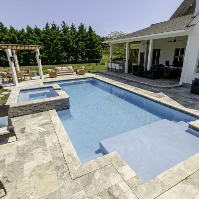 Backyard with a rectangular in-ground luxury pool, attached hot tub, stone patio, outdoor living areas, pergola, and a grass lawn bordered by tall trees.