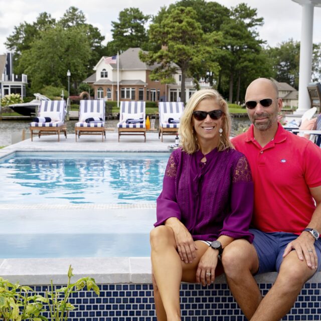 A man and woman sit side by side on the edge of a luxury pool, both wearing sunglasses. Lounge chairs, houses, trees, and a body of water are visible in the background, highlighting an inviting outdoor living space.