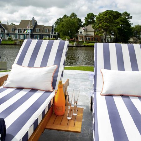 Two striped lounge chairs with white pillows face a waterfront, with a small table holding an orange bottle and two champagne glasses between them; perfect for relaxing after enjoying inground pools or custom spas. Houses and trees are visible across the water.
