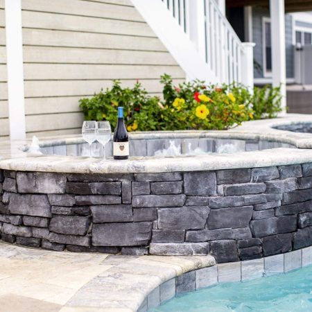 A stone hot tub next to an inground pool features a bottle of wine and two glasses on its edge; a flowering bush and house complete this inviting outdoor living scene.
