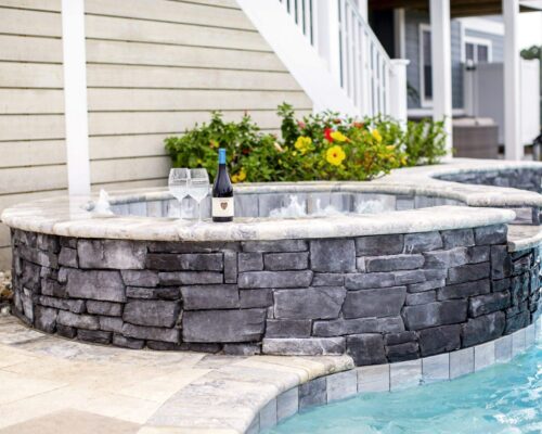 A stone hot tub next to an inground pool features a bottle of wine and two glasses on its edge; a flowering bush and house complete this inviting outdoor living scene.