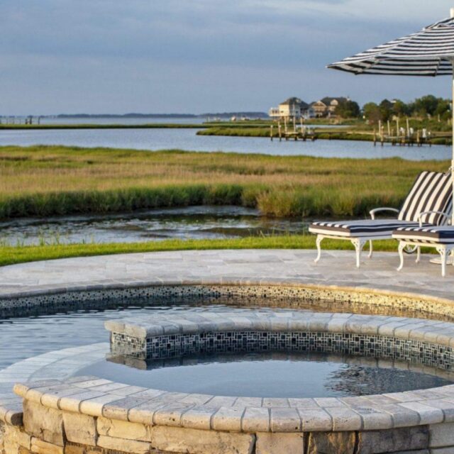 Stone-lined hot tub and pool with lounge chairs and striped umbrella, overlooking a grassy waterfront and distant houses under a cloudy sky—perfect for enjoying outdoor living.