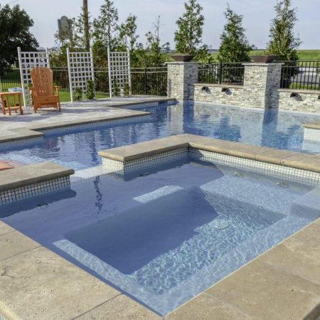 A modern backyard with a stone-tiled patio, hot tub in the foreground, luxury pool, lounge chairs, and a small table with drinks and towels.
