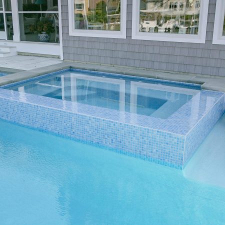 A tiled hot tub is built into a backyard fiberglass pool next to a modern house with large windows; a lounge chair floats in the pool, enhancing outdoor living.