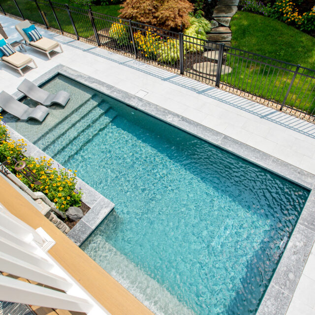 Rectangular inground pool with in-pool lounge chairs, steps, and a surrounding patio area with seating; bordered by a black fence and landscaped garden—perfect for those seeking luxury pools in their backyard retreat.
