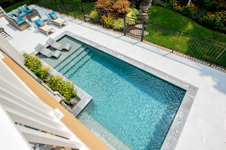 Rectangular inground pool with in-pool lounge chairs, steps, and a surrounding patio area with seating; bordered by a black fence and landscaped garden—perfect for those seeking luxury pools in their backyard retreat.