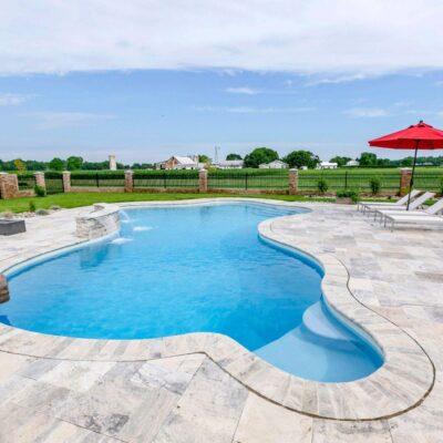 Outdoor swimming pool with clear water, surrounded by stone tiles and luxury pools features, lounge chairs with a red umbrella, plus a view of open fields and farm buildings in the background.
