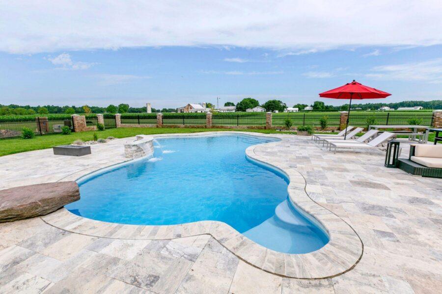 Outdoor swimming pool with clear water, surrounded by stone tiles and luxury pools features, lounge chairs with a red umbrella, plus a view of open fields and farm buildings in the background.