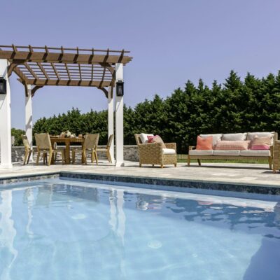 A backyard patio with a swimming pool, custom spas, wicker furniture, a pergola over a dining table, and a hedge of tall trees in the background under a clear sky.