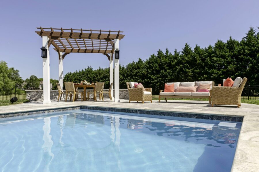 A backyard patio with a swimming pool, custom spas, wicker furniture, a pergola over a dining table, and a hedge of tall trees in the background under a clear sky.