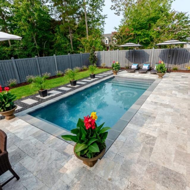 Rectangular backyard luxury pool with stone patio, lounge chairs, potted plants, and fenced yard bordered by trees creates the perfect outdoor living space.