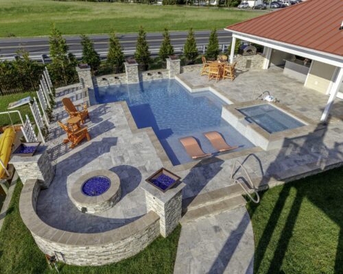 Aerial view of a backyard with an inground swimming pool, hot tub, lounge chairs, fire pit, outdoor living space, and a yellow slide near a grassy yard.