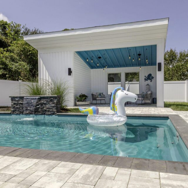 A backyard luxury pool with a stone waterfall feature, a large inflatable unicorn float, custom spa area, and a covered patio with seating under a blue ceiling.