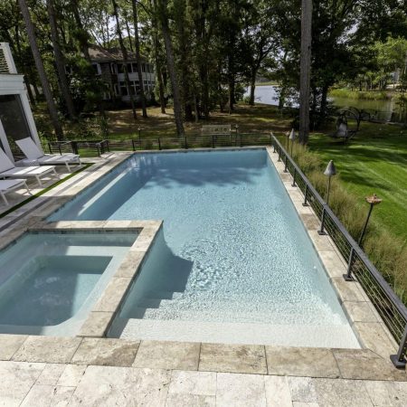 Rectangular backyard fiberglass pool with attached custom spa, surrounded by stone tiles, lounge chairs, metal railing, green lawn, trees, and a lake in the background.