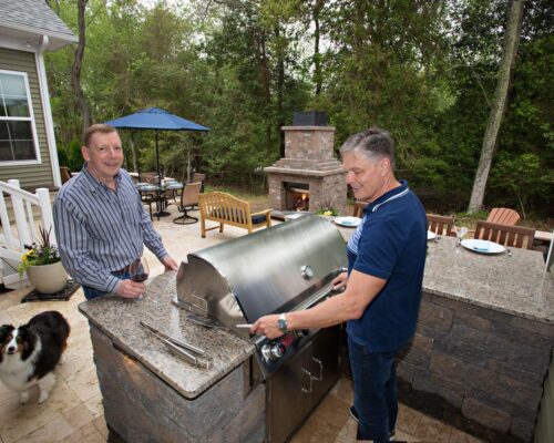 Two men stand by an outdoor grill on a patio, one grilling food and the other holding a wine glass. A dog is nearby, with an outdoor fireplace, luxury pools, and seating creating a perfect backdrop for relaxing evenings.