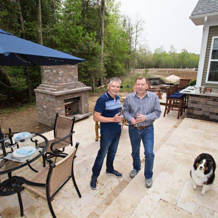 Two men stand on a stone patio with drinks, next to a dog. The patio features outdoor kitchens, a dining table, umbrella, grill, and outdoor fireplace, all set against a wooded background.