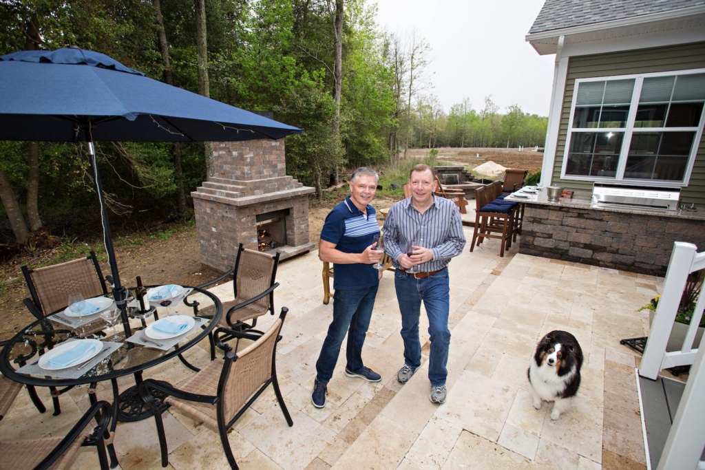 Two men stand on a patio near an outdoor kitchen with a grill and fireplace, holding drinks. A dog stands nearby, and a table is set for four. Trees and lawn are visible in the background.