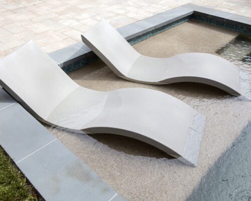 Two modern, curved lounge chairs partially submerged in shallow water beside a tiled fiberglass pool edge create the perfect setting for outdoor living on a sunny day.