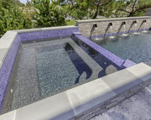 Rectangular outdoor inground pool with a raised custom spa section lined with blue tiles, stone coping, and water spouts along a stone wall.